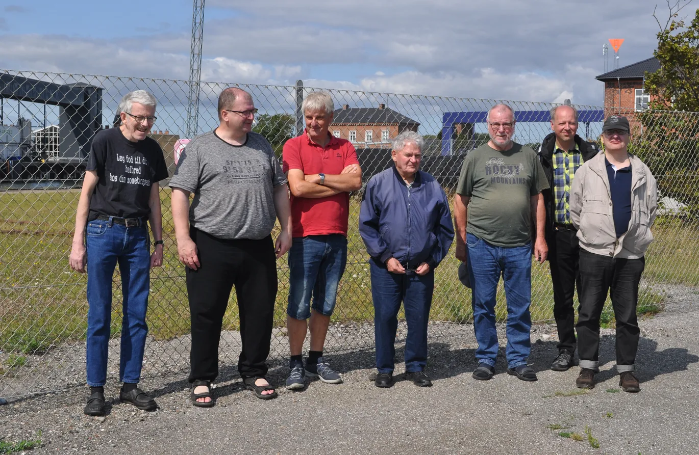 Sommerudflugt til Gedser Remise<br>Fra venstre: Thorben Rostgaard, Per Vad Nielsen, Uffe Bek Pedersen, Jan Hally. Mogens Christensen, Andreas Fischer og Artur Frenzel.<br> Yderst til venstre ses lidt af færgehallen fra 1903, og yderst til højre ses et hjørne af Gedsers oprindelige stationsbygning fra 1886.